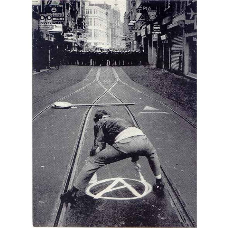 Prentbriefkaart van een man die het logo van anarchisme op straat schildert, met verderop een cordon politieagenten